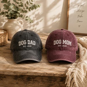 Könnte beinhalten: Zwei Baseballkappen mit verwaschener Optik. Die linke Kappe ist anthrazitfarben mit weißem Sticktext DOG DAD EST. 2026. Die rechte Kappe ist kastanienbraun mit weißem Sticktext DOG MOM EST. 2026. Beide Kappen haben einen gebogenen Schirm.