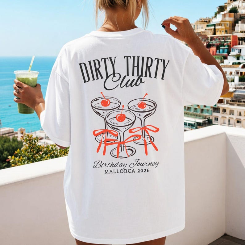 May include: A white t-shirt featuring a black and red graphic on the back. The text reads DIRTY THIRTY Club, Birthday Journey, and MALLORCA 2026. The design includes three martini glasses with red cherries and red ribbons tied around the stems.