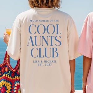 May include: Cream-colored t-shirt with the text "PROUD MEMBER OF THE COOL AUNTS CLUB" in blue. The shirt also includes the names "LISA & MICHAEL" and the year "EST. 2027". The shirt is part of a club-themed collection.