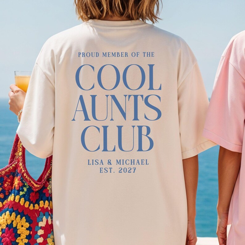 May include: Cream-colored t-shirt with the text "PROUD MEMBER OF THE COOL AUNTS CLUB" in blue. The shirt also includes the names "LISA & MICHAEL" and the year "EST. 2027". The shirt is part of a club-themed collection.