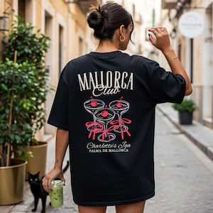 May include: A black t-shirt featuring a graphic on the back with the text "MALLORCA Club" above three cocktail glasses, each garnished with a red cherry and tied with a pink ribbon. Below the glasses, the text reads "Charlotte's Iga PALMA DE MALLORCA."