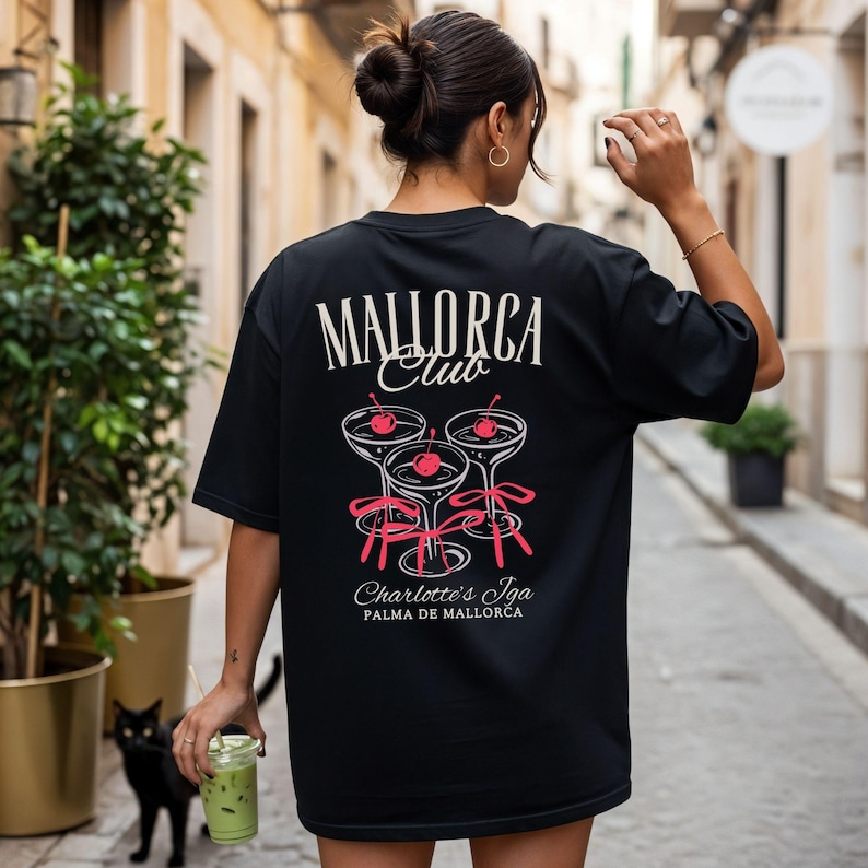 May include: A black t-shirt featuring a graphic on the back with the text "MALLORCA Club" above three cocktail glasses, each garnished with a red cherry and tied with a pink ribbon. Below the glasses, the text reads "Charlotte's Iga PALMA DE MALLORCA."