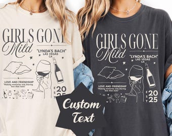 May include: Two t-shirts, one cream and one charcoal, feature the text "GIRLS GONE Mild" with "LYNDA'S BACH" in Las Vegas, CA. The design includes illustrations of a wine glass, bottle, and other elements. The shirts also have the text "LOVE AND FRIENDSHIP".