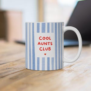 Könnte beinhalten: Ein weißer Keramikbecher mit hellblauen vertikalen Streifen und einem zentralen weißen rechteckigen Feld. Der Text COOL AUNTS CLUB ist in roten Großbuchstaben auf das Feld gedruckt, ergänzt durch ein kleines rotes Herz unter dem Text.