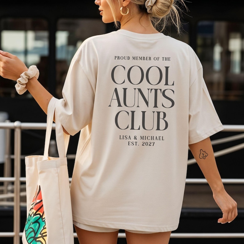 May include: Cream-colored t-shirt with the text "PROUD MEMBER OF THE COOL AUNTS CLUB" in dark gray. The text "LISA & MICHAEL EST. 2027" is below. The shirt is part of a casual wear collection.