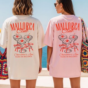 May include: Two oversized t-shirts, one cream and one light pink, featuring a red graphic on the back. The design displays the text "MALLORCA Club" above three cocktail glasses with cherries and red ribbons, and "Charlotte's Iga PALMA DE MALLORCA" below.