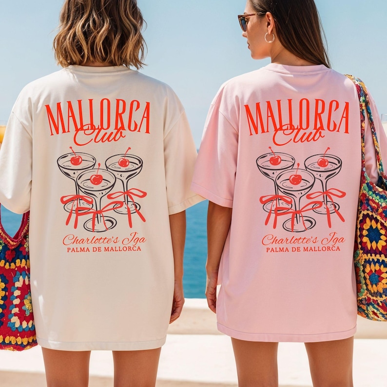 May include: Two oversized t-shirts, one cream and one light pink, featuring a red graphic on the back. The design displays the text "MALLORCA Club" above three cocktail glasses with cherries and red ribbons, and "Charlotte's Iga PALMA DE MALLORCA" below.