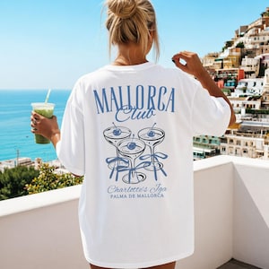 May include: A white short-sleeved t-shirt featuring a blue graphic on the back. The design includes the text "MALLORCA Club" above three cocktail glasses with cherries and ribbons, with the text "Charlotte's Iga PALMA DE MALLORCA" below.