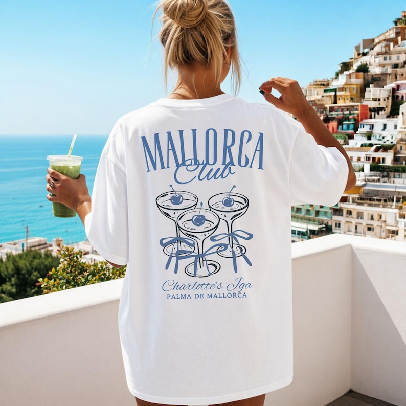 May include: A white short-sleeved t-shirt featuring a blue graphic on the back. The design includes the text "MALLORCA Club" above three cocktail glasses with cherries and ribbons, with the text "Charlotte's Iga PALMA DE MALLORCA" below.