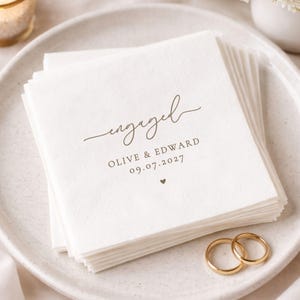 May include: A stack of off-white paper napkins with the word "engaged" in a cursive font. Below the word are the names "OLIVE & EDWARD" and the date "09.07.2027". Two gold wedding rings are on the plate.