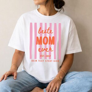 May include: White t-shirt with a pink and orange design. The design includes pink vertical stripes and the words "beste MOM ever" in orange, with "EST. 2017" below. The text "DEIN TEXT STEHT HIER" is at the bottom.