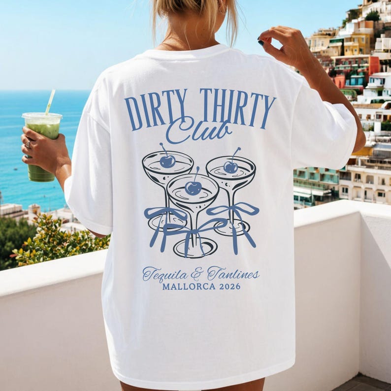 May include: A white t-shirt featuring a blue graphic on the back with the text "DIRTY THIRTY Club" above three illustrated cocktail glasses, each containing a cherry and tied with a bow. Below the glasses is the text "Tequila & Tanlines MALLORCA 2026".