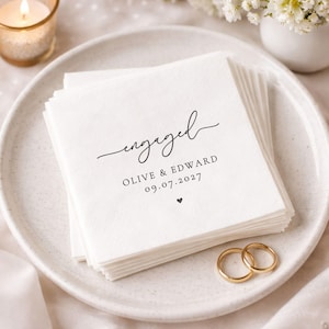 May include: A stack of white paper napkins with the word "engaged" in elegant script. Below the word are the names "Olive & Edward" and the date "09.07.2027". A small heart is printed below the date. Two gold wedding rings are on the plate.