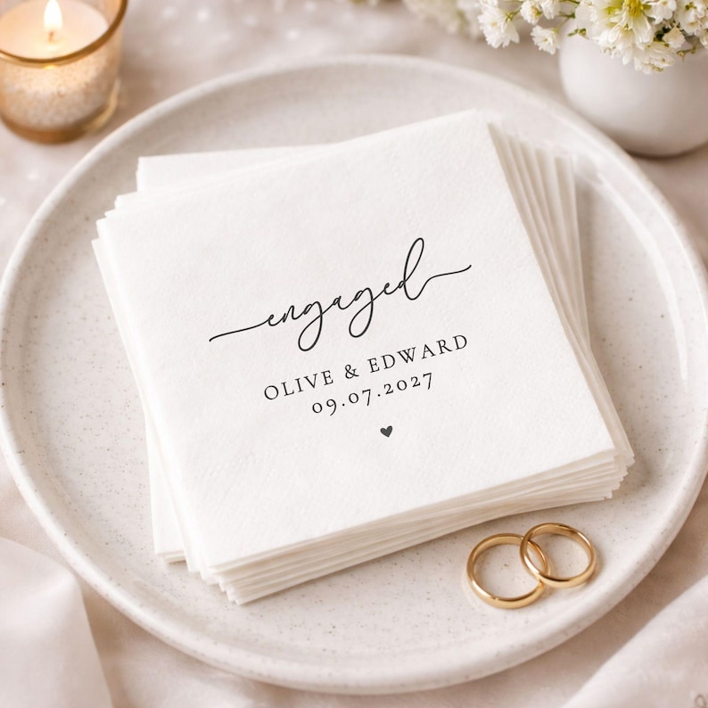 May include: A stack of white paper napkins with the word "engaged" in elegant script. Below the word are the names "Olive & Edward" and the date "09.07.2027". A small heart is printed below the date. Two gold wedding rings are on the plate.