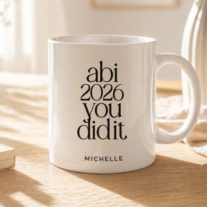 K&ouml;nnte beinhalten: Ein wei&szlig;er Keramikbecher mit schwarzem Text, der "abi 2026 you did it" in einer gestapelten Serifenschrift zeigt. Darunter ist der Name "MICHELLE" in einer kleineren serifenlosen Schrift gedruckt. Der Becher hat einen Henkel und eine glatte Oberfl&auml;che.