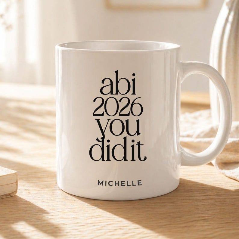 K&ouml;nnte beinhalten: Ein wei&szlig;er Keramikbecher mit schwarzem Text, der "abi 2026 you did it" in einer gestapelten Serifenschrift zeigt. Darunter ist der Name "MICHELLE" in einer kleineren serifenlosen Schrift gedruckt. Der Becher hat einen Henkel und eine glatte Oberfl&auml;che.