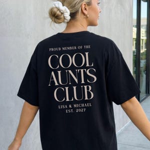 May include: Black t-shirt with the text "PROUD MEMBER OF THE COOL AUNTS CLUB" in a serif font. Below the text are the names "LISA & MICHAEL" and the year "EST. 2027". The shirt is worn by a person with blonde hair.