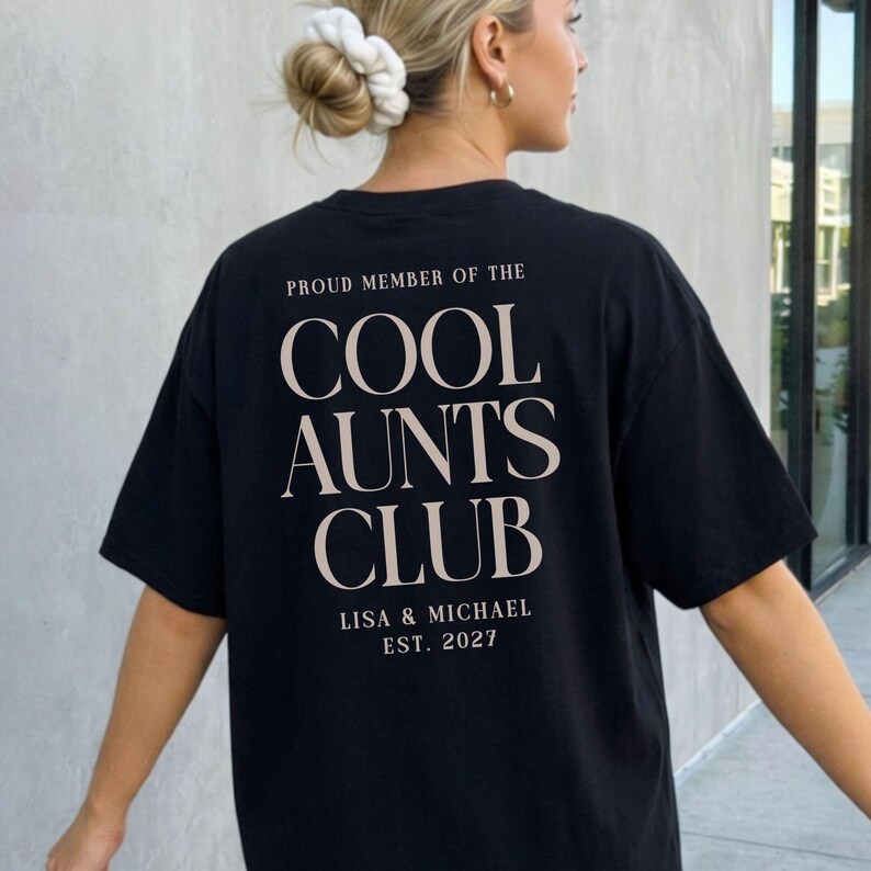 May include: Black t-shirt with the text "PROUD MEMBER OF THE COOL AUNTS CLUB" in a serif font. Below the text are the names "LISA & MICHAEL" and the year "EST. 2027". The shirt is worn by a person with blonde hair.