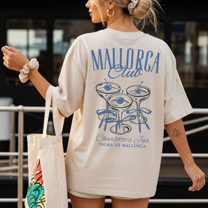 May include: An oversized cream-colored t-shirt featuring a blue graphic on the back. The design displays three cocktail glasses with cherries and ribbons, with the text "MALLORCA Club", "Charlotte's Iga", and "PALMA DE MALLORCA" printed in blue.