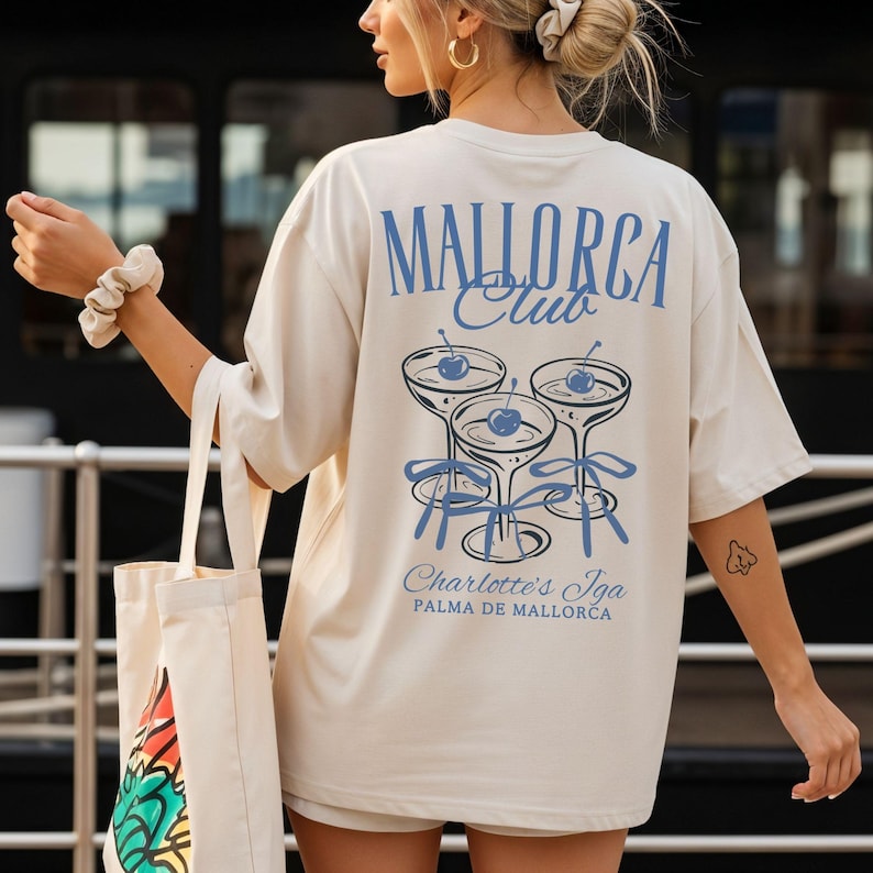 May include: An oversized cream-colored t-shirt featuring a blue graphic on the back. The design displays three cocktail glasses with cherries and ribbons, with the text "MALLORCA Club", "Charlotte's Iga", and "PALMA DE MALLORCA" printed in blue.