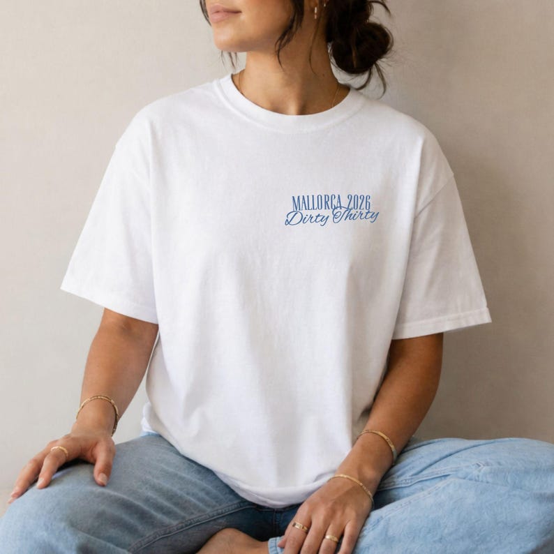 May include: A white short-sleeved t-shirt featuring blue text on the upper left chest area. The text reads Mallorca 2026 in a block font, with the words Dirty Thirty written underneath in a cursive script style. The shirt has a crew neckline.