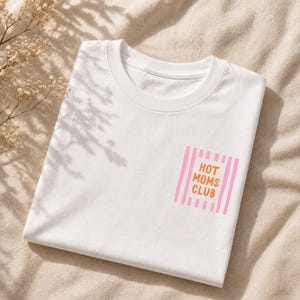 Könnte beinhalten: Ein weißes Kurzarm-T-Shirt mit einer rechteckigen Grafik auf der Brust. Das Design besteht aus vertikalen hellrosa Streifen mit dem orangefarbenen Text HOT MOMS CLUB in der Mitte. Das T-Shirt hat einen klassischen Rundhalsausschnitt.