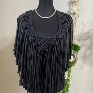 Puede incluir: Un top negro con flecos, un escote de ganchillo y un collar de perlas. El top se exhibe en un maniquí negro. Los flecos cuelgan de los hombros y el pecho. El top es una prenda de vestir única.
