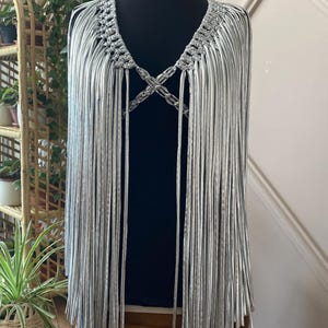 以下が含まれることがあります： 織り込まれたネックラインとクロスディテールが特徴のシルバーフリンジケープ。黒いマネキンに飾られています。フリンジが垂れ下がり、レイヤード効果を生み出しています。このケープは、ステートメントピースです。