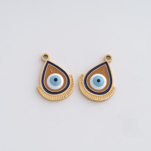 Op de afbeelding: Twee goudkleurige druppelvormige bedels met een boze oog-ontwerp. Elke bedel heeft een lichtblauw oog met een zwarte pupil, omgeven door een witte cirkel, een bruin achtergrond en een donkerblauwe rand. De bedels hebben een kleine lus aan de bovenkant om op te hangen.