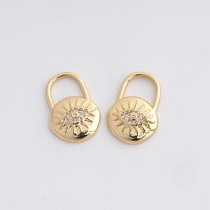 May include: Two gold-colored padlock charms. Each charm features an eye design with a central cluster of small, clear crystals. The padlocks have a rounded shape with a curved handle at the top, and are set against a white background.