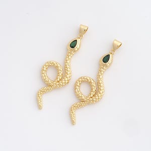Peut inclure: Deux pendentifs dorés en forme de serpent avec des yeux en pierres précieuses vertes. Les pendentifs ont un motif texturé en forme d'écailles et une boucle en haut pour une chaîne. Les corps des serpents sont enroulés.