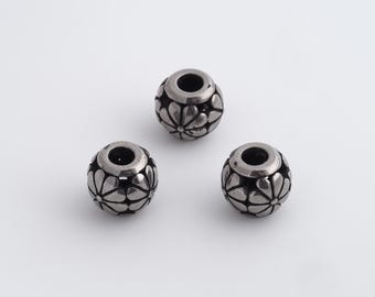 Puede incluir: Tres cuentas plateadas con un diseño floral. Cada cuenta tiene un centro oscuro y está decorada con un patrón de pétalos estilizados. Las cuentas son esféricas y tienen una superficie lisa y reflectante.