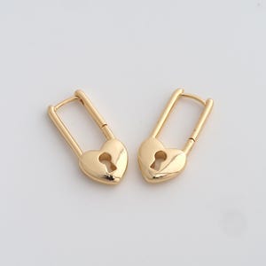 May include: A pair of gold-colored heart-shaped earrings. Each earring features a keyhole design and a hinged clasp. The earrings are displayed on a white background.