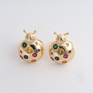Könnte beinhalten: Ein Paar goldfarbene Marienkäfer-Ringe. Jeder Ring hat einen abgerundeten Körper, der mit farbigen Edelsteinen verziert ist, darunter Grün, Blau, Rosa und Gelb. Der Kopf jedes Marienkäfers ist mit kleinen, klaren Steinen und Fühlern verziert.
