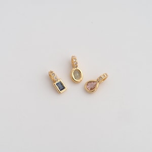 Peut inclure: Trois pendentifs dorés avec des pierres précieuses de formes différentes : une pierre bleue rectangulaire, une pierre verte ovale et une pierre rose en forme de goutte. Chaque pendentif a une petite boucle ornée de petites pierres claires.