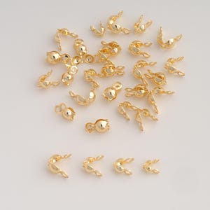 May include: A collection of gold-toned metal crimp covers, also known as bead tips, scattered across the frame. Each piece features a small, rounded, hollow cup shape with a tiny loop at the top, designed for securing jewelry wire ends.