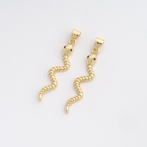 Puede incluir: Dos colgantes dorados con forma de serpiente y ojos verdes. Los colgantes presentan un patrón texturizado en forma de escamas. Cada serpiente tiene un lazo en la parte superior para sujetarlo a una cadena. El fondo es blanco liso.