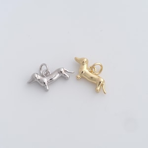 May include: Two dachshund dog charms, one silver and one gold, are shown against a white backdrop. The charms are small, detailed, and have a loop at the top for attaching to a necklace or bracelet. The dogs are in a standing position.