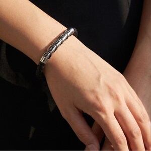 Könnte beinhalten: Ein Armband mit einem geflochtenen schwarzen Lederband, das mit einer Reihe polierter, silberfarbener zylindrischer Metallperlen verziert ist. Jede Metallperle weist ein sich wiederholendes eingraviertes Muster auf, das einen strukturierten Kontrast zum dunklen Lederband bildet.