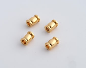 Könnte beinhalten: Vier kleine, goldfarbene Metallperlen sind auf einer weißen Oberfläche angeordnet. Jede Perle hat eine zylindrische Form mit einer leicht breiteren Mitte und einem Loch in der Mitte. Die Perlen haben eine glänzende, polierte Oberfläche.