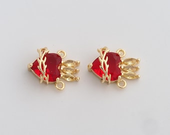 May include: Two gold-colored heart-shaped charms with red gemstone centers and yellow-gold accents. Each charm has a loop for attaching to jewelry. The charms are set against a plain white background.