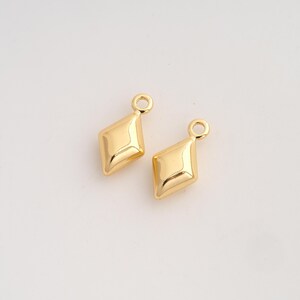May include: Two small, polished gold-tone metal charms in a diamond or rhombus shape. Each charm features a smooth, faceted surface with a single circular jump ring attached at the top for hanging, reflecting light with a bright, metallic finish.
