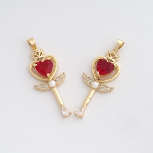 May include: Two gold-colored pendants with a heart-shaped red gemstone, a crown, and small wings. Each pendant has a white pearl and a teardrop-shaped clear gemstone at the bottom. The pendants are on a white background.