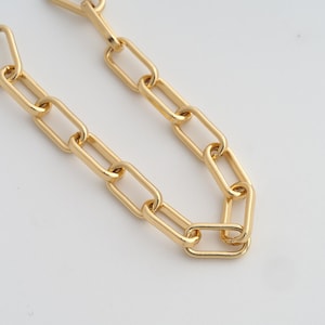 Op de afbeelding: Close-up van een goudkleurige ketting met grote, rechthoekige schakels. De ketting is gemaakt van een glanzend, metalen materiaal en wordt gepresenteerd tegen een witte achtergrond. De schakels zijn gelijkmatig verdeeld en creëren een gedurfde, moderne uitstraling.