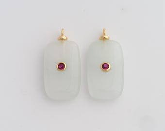 Puede incluir: Dos colgantes rectangulares con bordes redondeados. Cada colgante es de un verde pálido y translúcido con una pequeña gema roja y redonda en el centro. Los colgantes tienen lazos dorados en la parte superior para colgar.