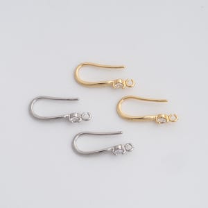 May include: Four earring hooks are shown, two gold-coloured and two silver-coloured, against a white backdrop. Each hook has a curved design with a small, clear gemstone near the loop. These hooks are for jewellery making.