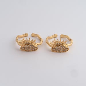 May include: Two gold-colored adjustable rings. Each ring features a sun and cloud design. The sun has pointed rays, and the cloud is encrusted with small, clear stones. The rings are on a white background.