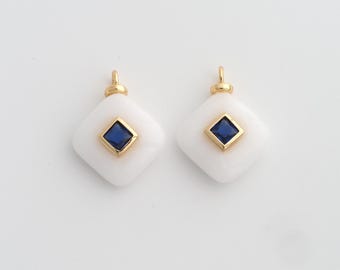 May include: Two white, diamond-shaped pendants. Each has a gold border and a square, dark blue gemstone in the centre. A small gold loop is at the top of each pendant for hanging. The background is plain white.