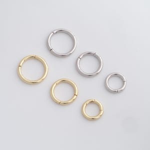 May include: Six hoop earrings in gold and silver tones are arranged on a white surface. The earrings vary in size, with three gold and three silver hoops. The hoops have a hinged design for easy wear.