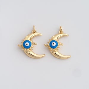 May include: Two gold-colored crescent moon charms with a sunburst design and a blue and white eye in the center. The charms have a loop at the top for hanging and are set against a white background.